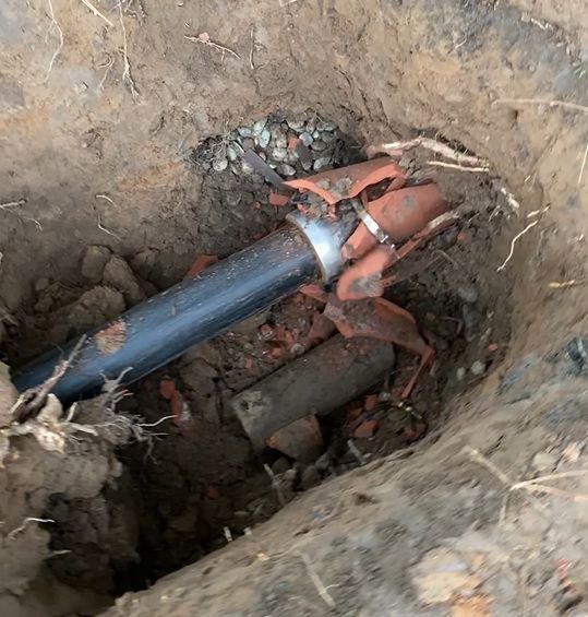 Broken underground pipes in a dirt hole, showing a black pipe connected to broken orange and gray pipes.
