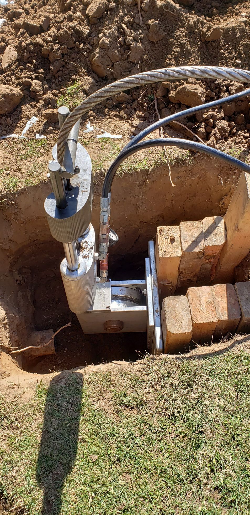 A cable-pulling machine in a dug-out hole, with a metal cable and hydraulic hoses, set on grass and dirt.