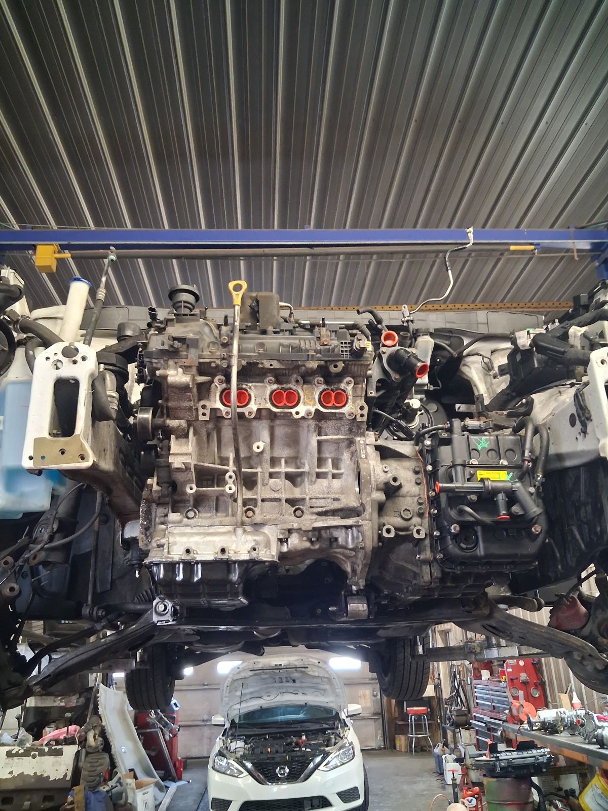 Car engine suspended in a workshop, with tools and a white car below. | Kensinger Repair Center