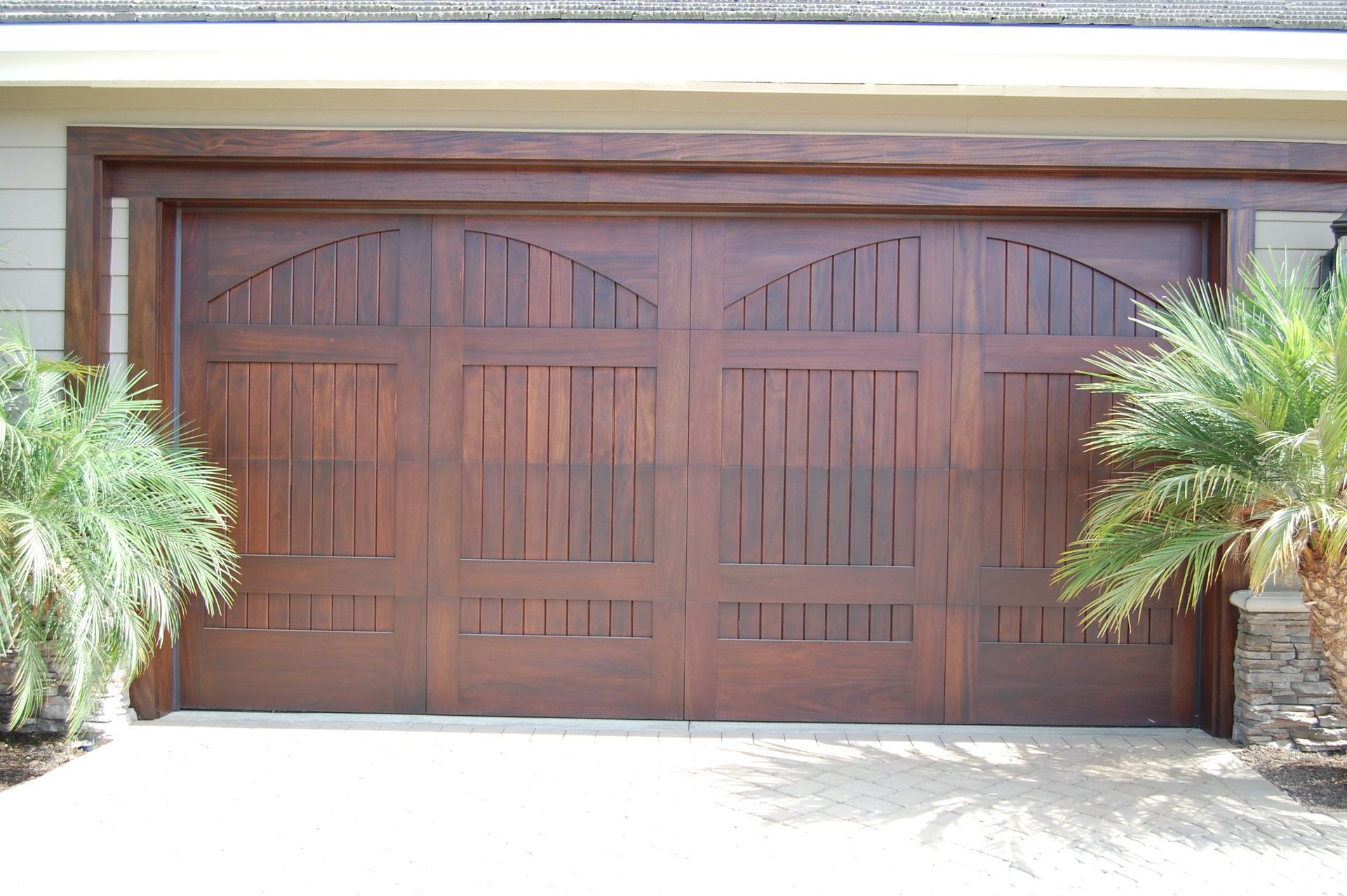 Wooden double garage doors with arched top panels, framed by stone and palm plants.