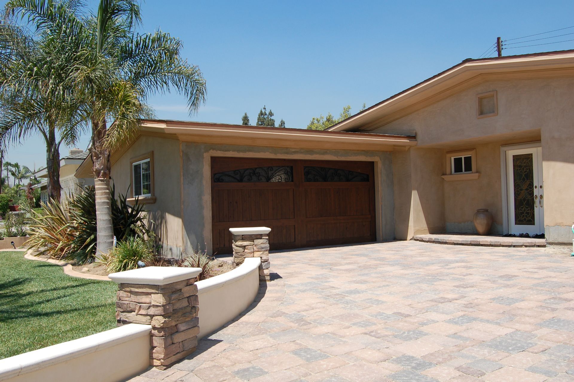 Single-story beige house with a garage, palm tree, and stone-paved driveway under a clear sky