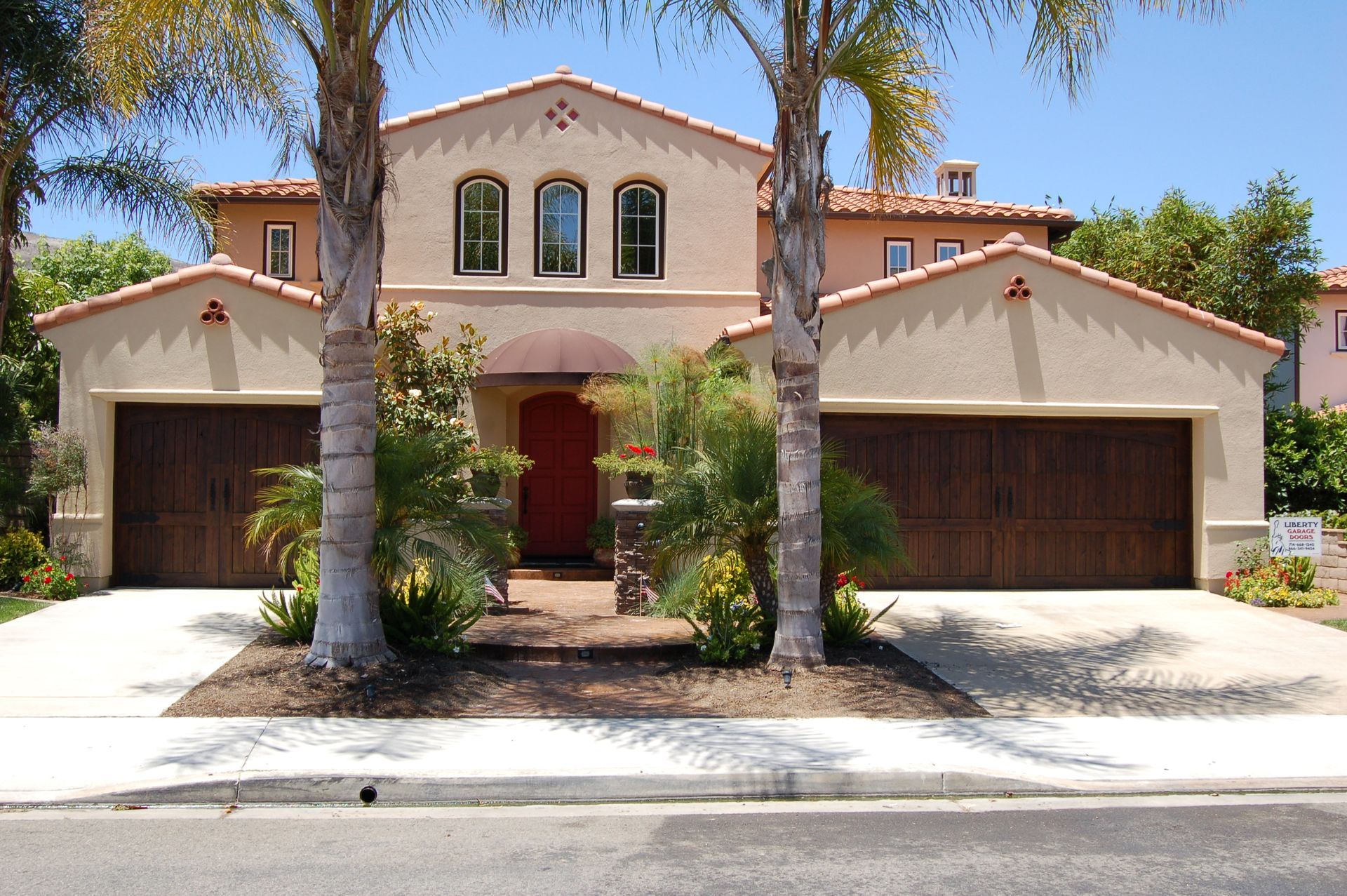 Two-story stucco house with red front door, arched windows, palm trees, and a two-car garage