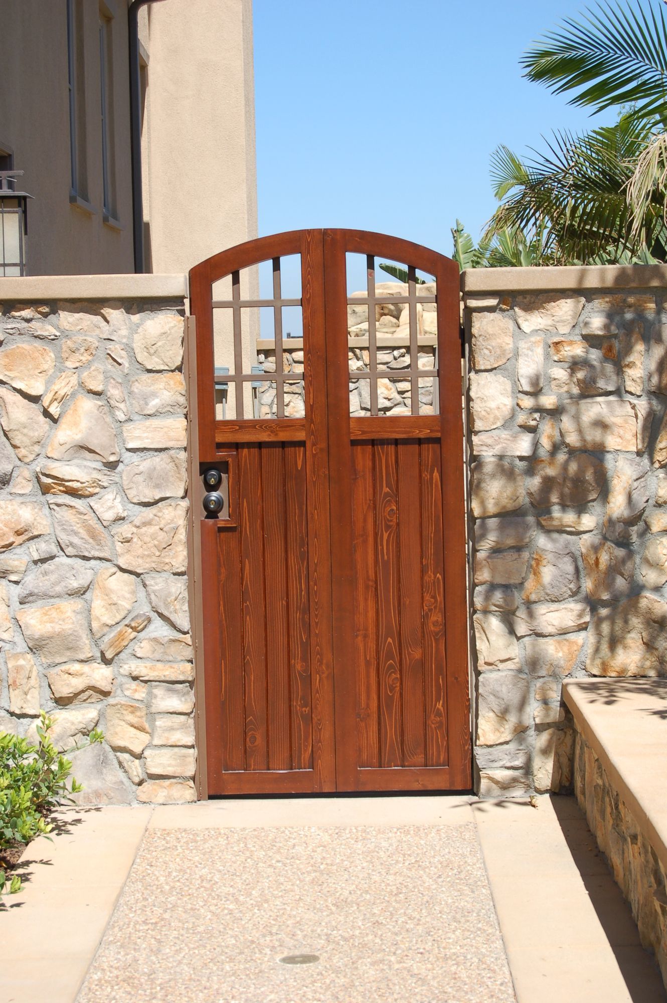 Wooden gate in a stone wall opening onto a sunlit pathway, with a palm tree nearby.