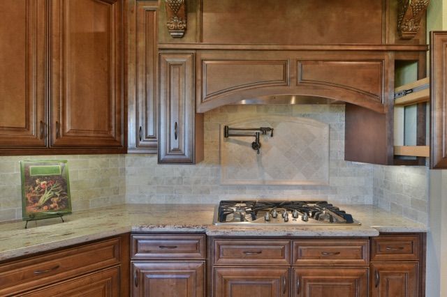 Wooden kitchen with beige tile backsplash, granite counters, and a stainless gas cooktop beneath an arched hood.