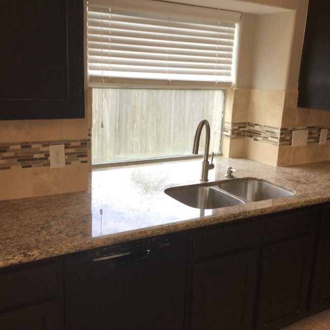 Kitchen countertop with stainless sink beneath a window and white blinds, dark cabinets, and tiled backsplash.
