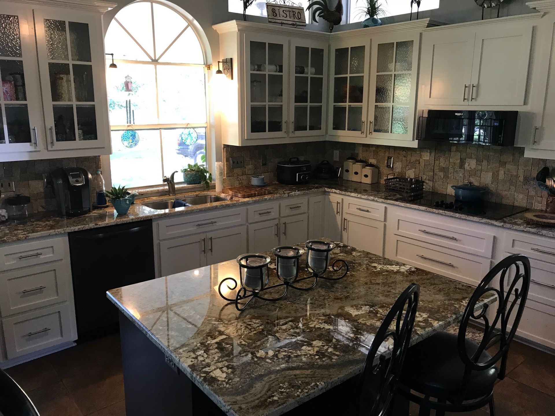 Bright kitchen with granite island, white cabinets, large arched window, and bar stools.