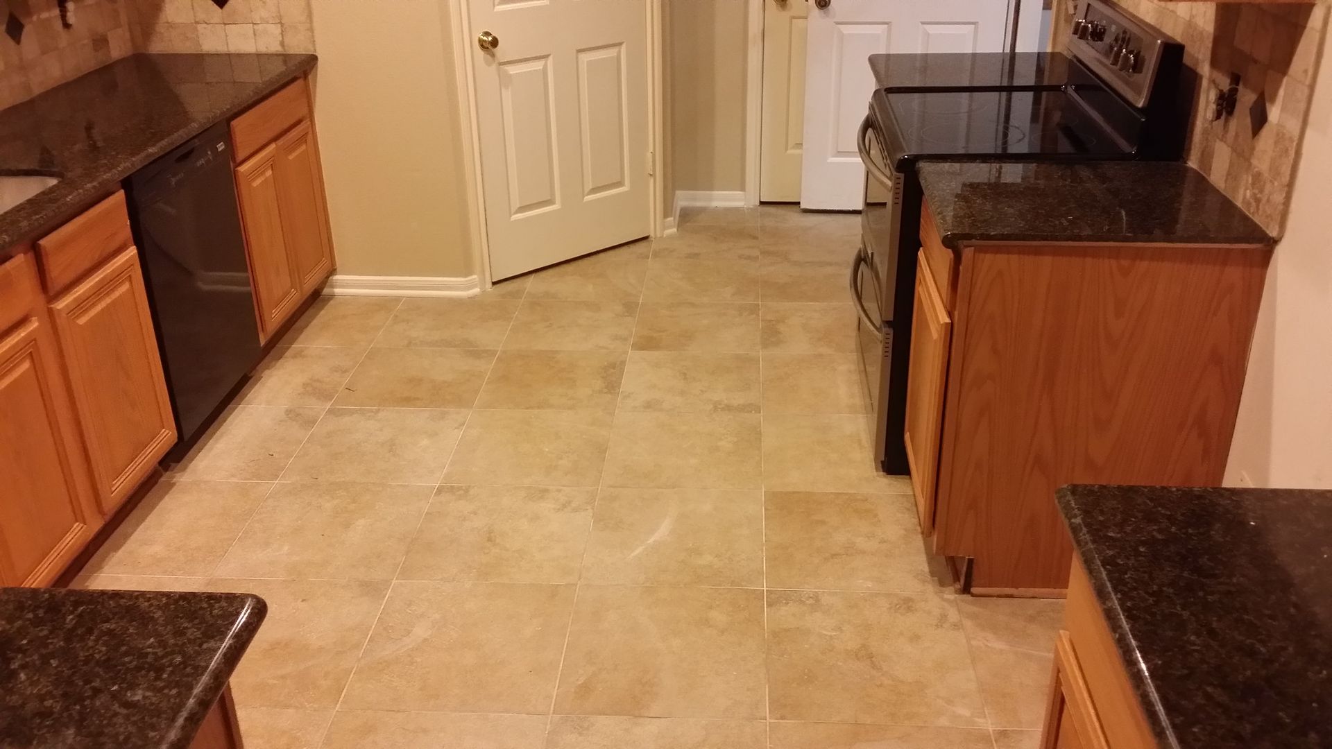 Empty kitchen with beige tile floor, wooden cabinets, and dark granite countertops.