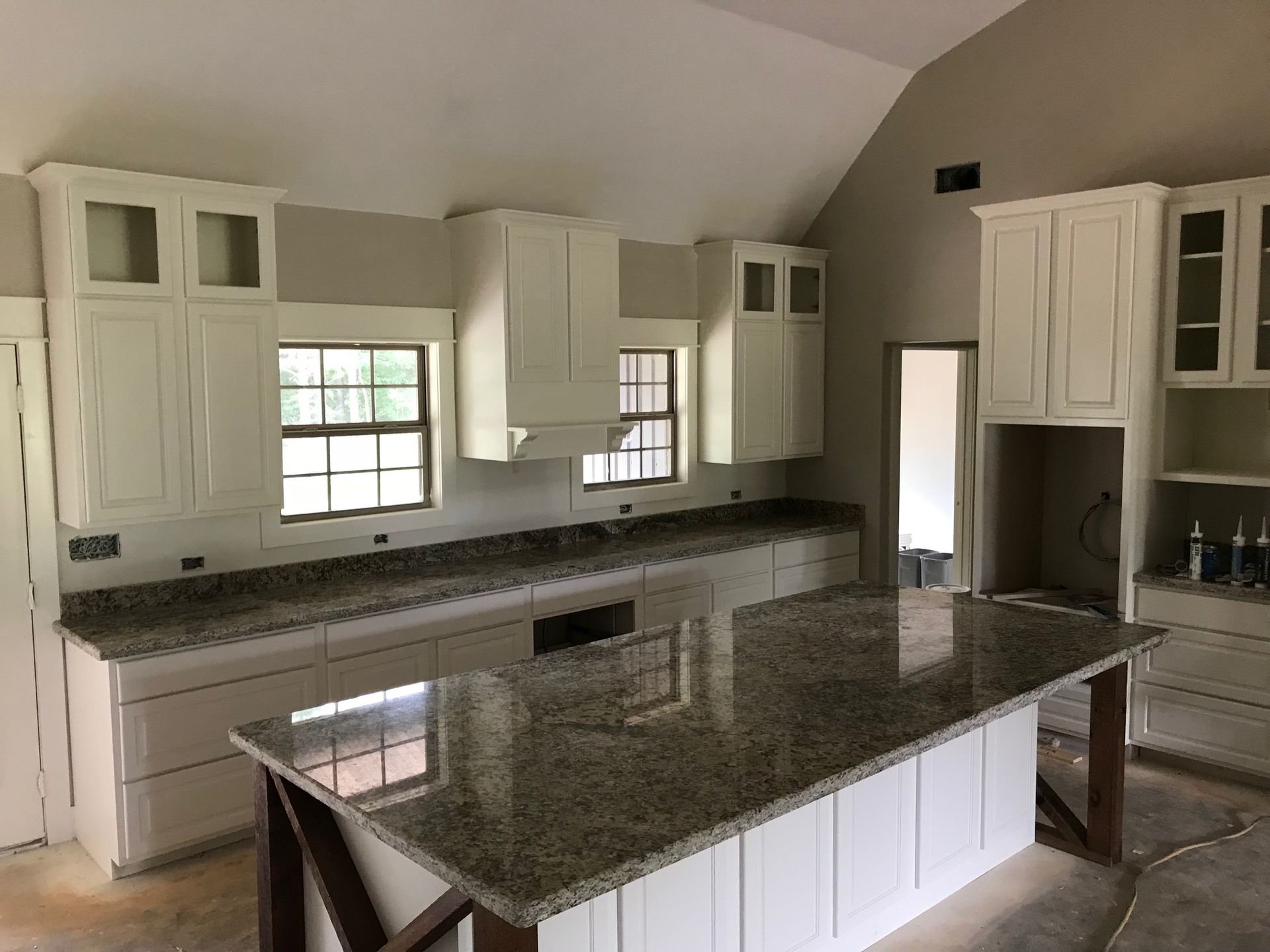 Bright kitchen with white cabinets, dark granite island, and large windows under a vaulted ceiling