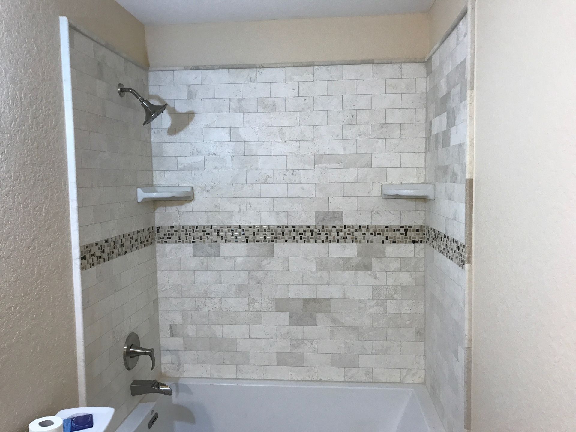 Empty tiled bathtub/shower with white and gray tile surround and chrome fixtures.