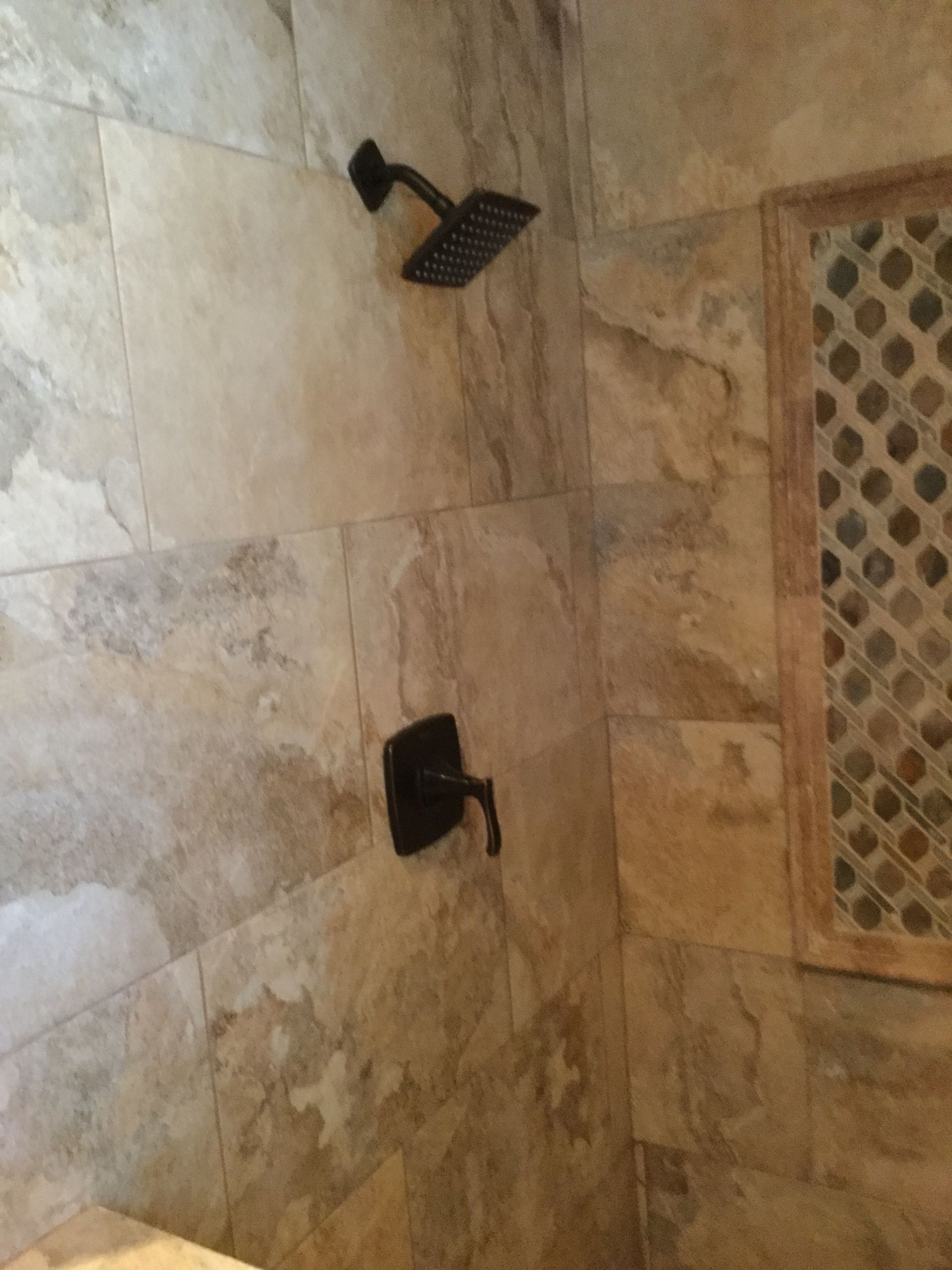 Two black shower fixtures on beige tiled bathroom wall beside a small window grate