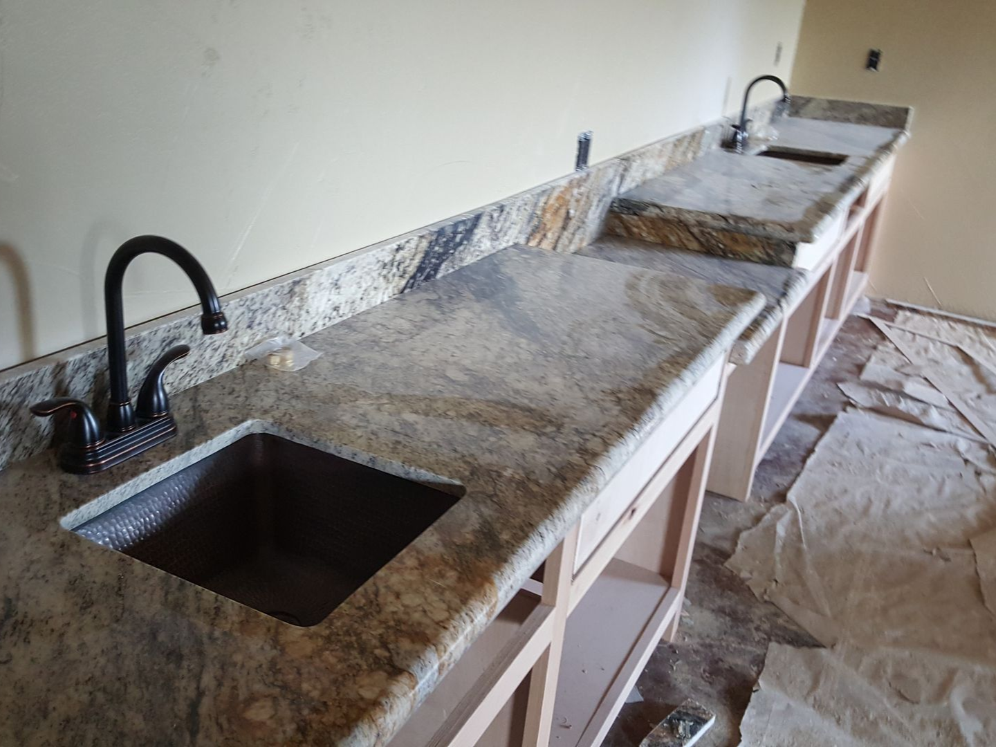 Long granite countertop with two black sinks and faucets, covered in protective plastic during kitchen installation