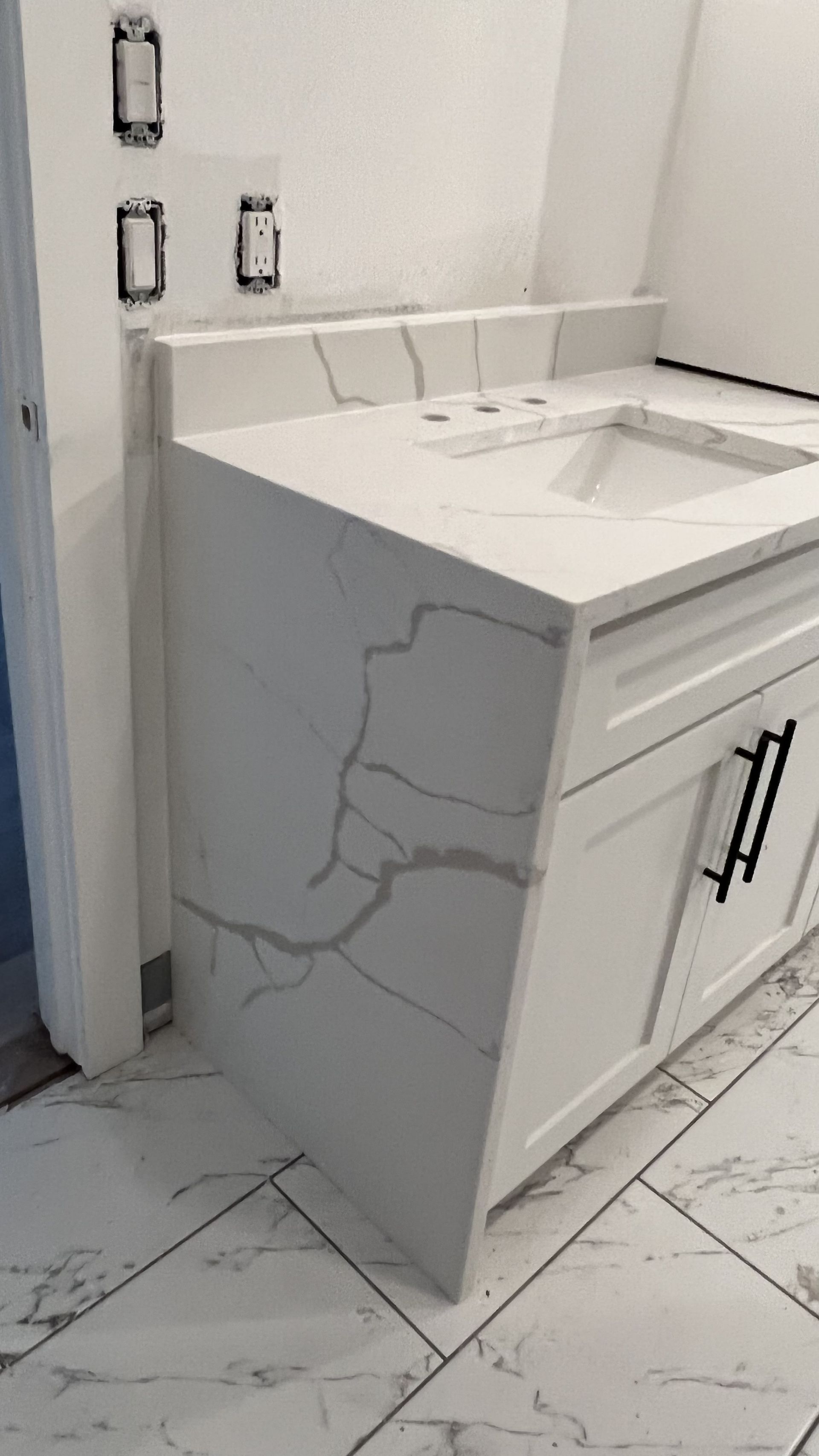 White bathroom vanity with sink, marble-look cabinet, and cracked side panel on tiled floor.