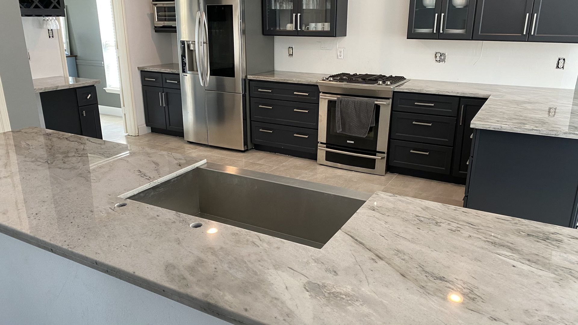 Modern kitchen with gray island and sink, stainless appliances, dark cabinets, and marble countertops.