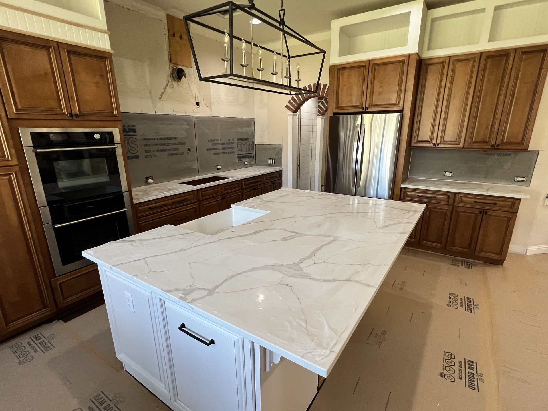 Bright kitchen with large white island, wood cabinets, built-in ovens, and hanging light fixture.