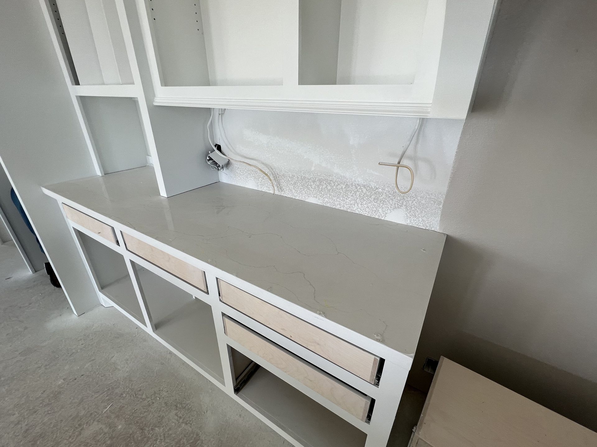 White built-in desk with drawers and shelves in an empty room, with carpeted floor and wall outlet area behind it