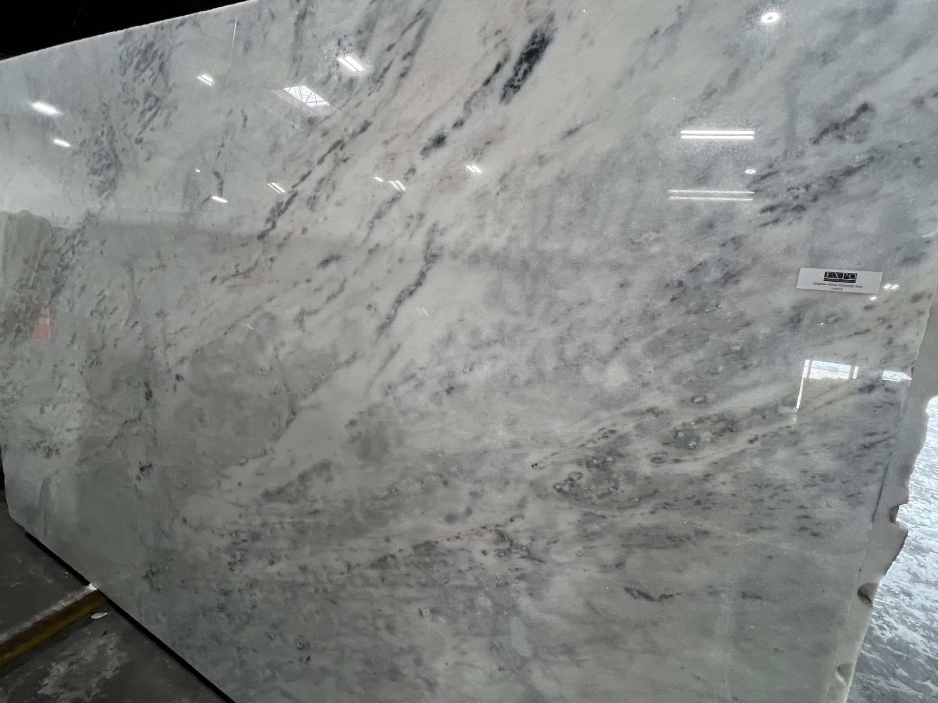 Large polished white-and-gray marble slab with subtle veining, shown upright in a warehouse or showroom.