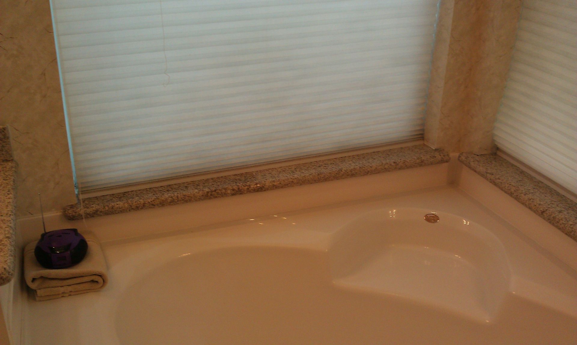 White corner bathtub under two beige window shades with a small soap dish and dark object on the left ledge