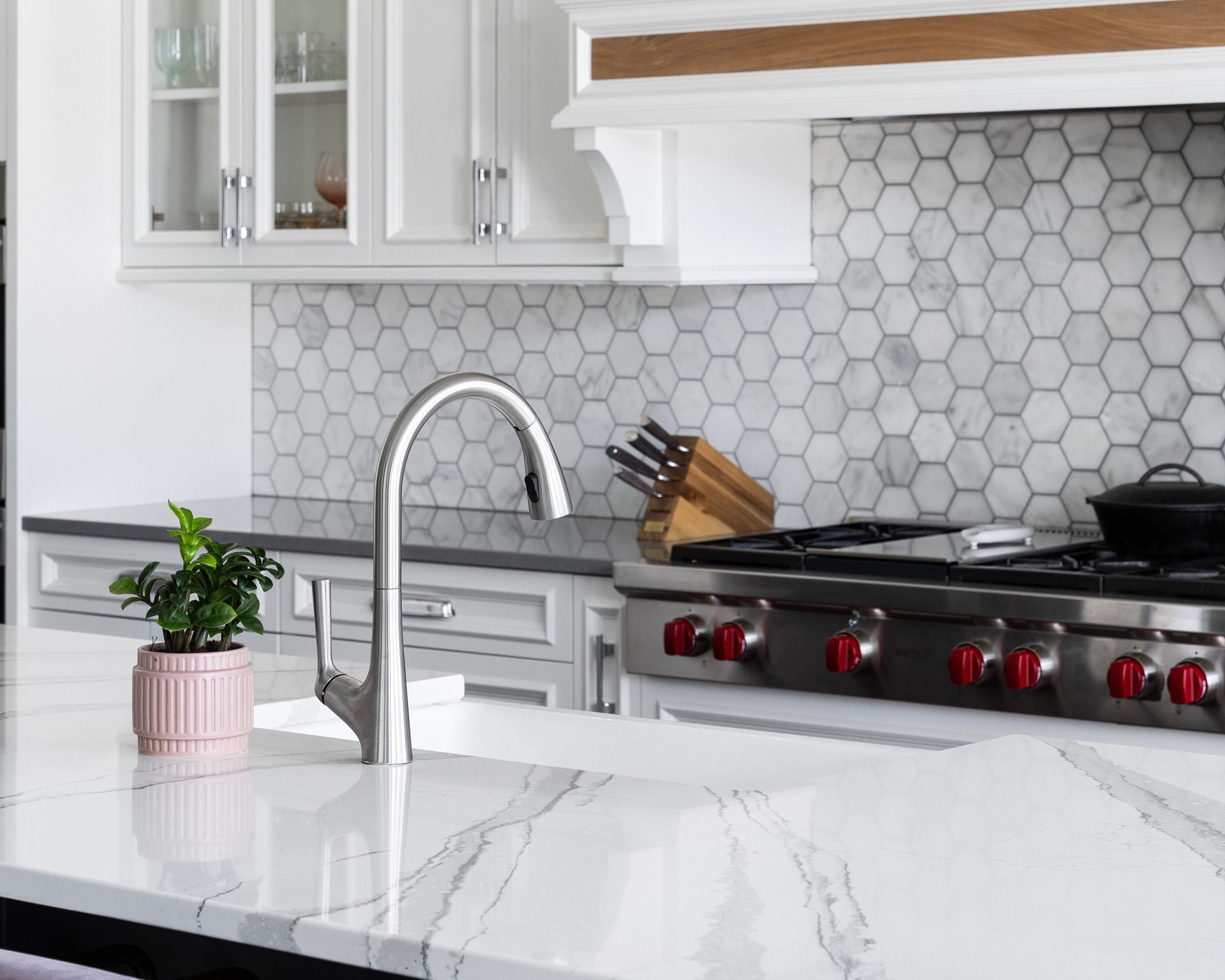 Bright white kitchen with marble island, stainless stove, hex tile backsplash, and a potted plant by the sink