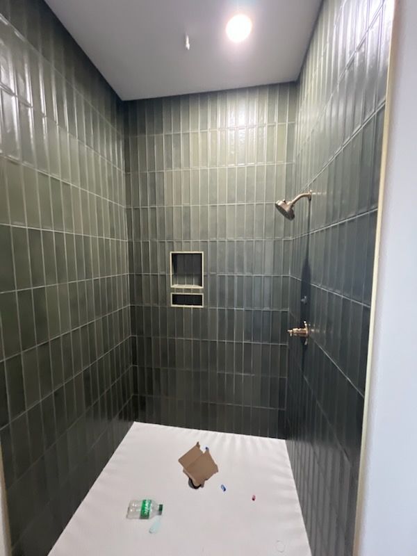 Empty tiled shower stall with white floor, green-gray walls, showerhead, niche, and a cardboard box on the floor