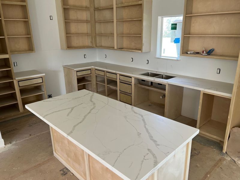 Unfinished kitchen with white countertops, wood cabinets, and a marble-look island in the center.
