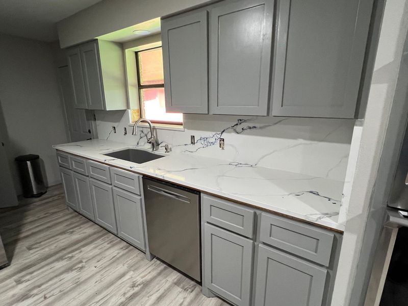 Modern gray kitchen with white marble countertops, stainless dishwasher, and a window over the sink