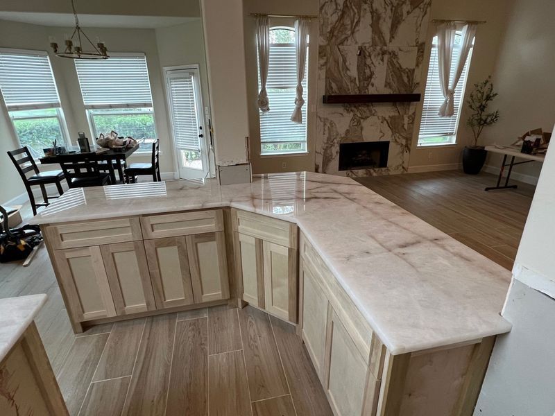 Empty kitchen with light wood cabinets, marble island, and open view to a dining area and beach beyond.