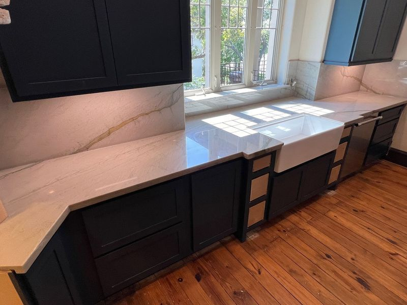 Modern kitchen with black cabinets, white marble countertops, and a large window over a wood floor