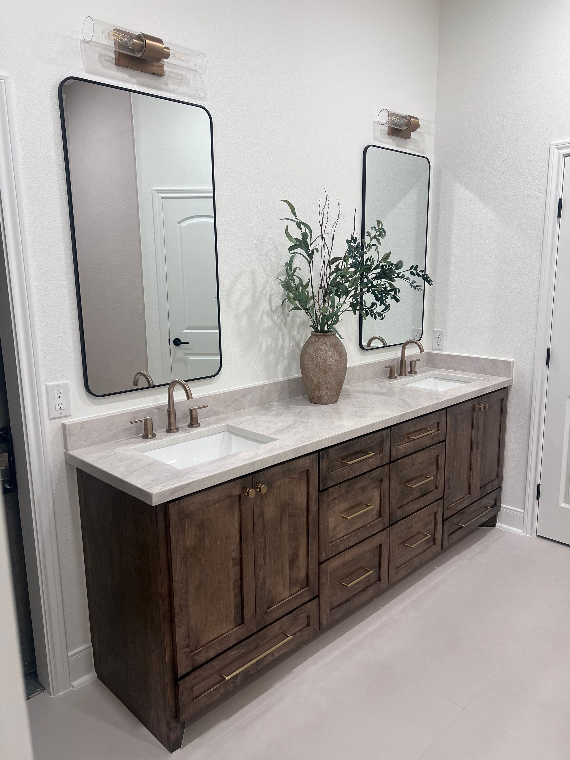 Double vanity bathroom with wood cabinets, white countertops, mirrors, and a vase of greenery.