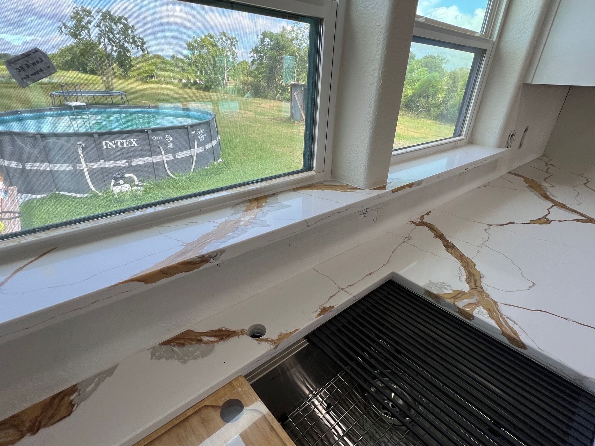 Damaged windowsill with peeling paint and cracks above a floor vent, overlooking a yard and above-ground pool.