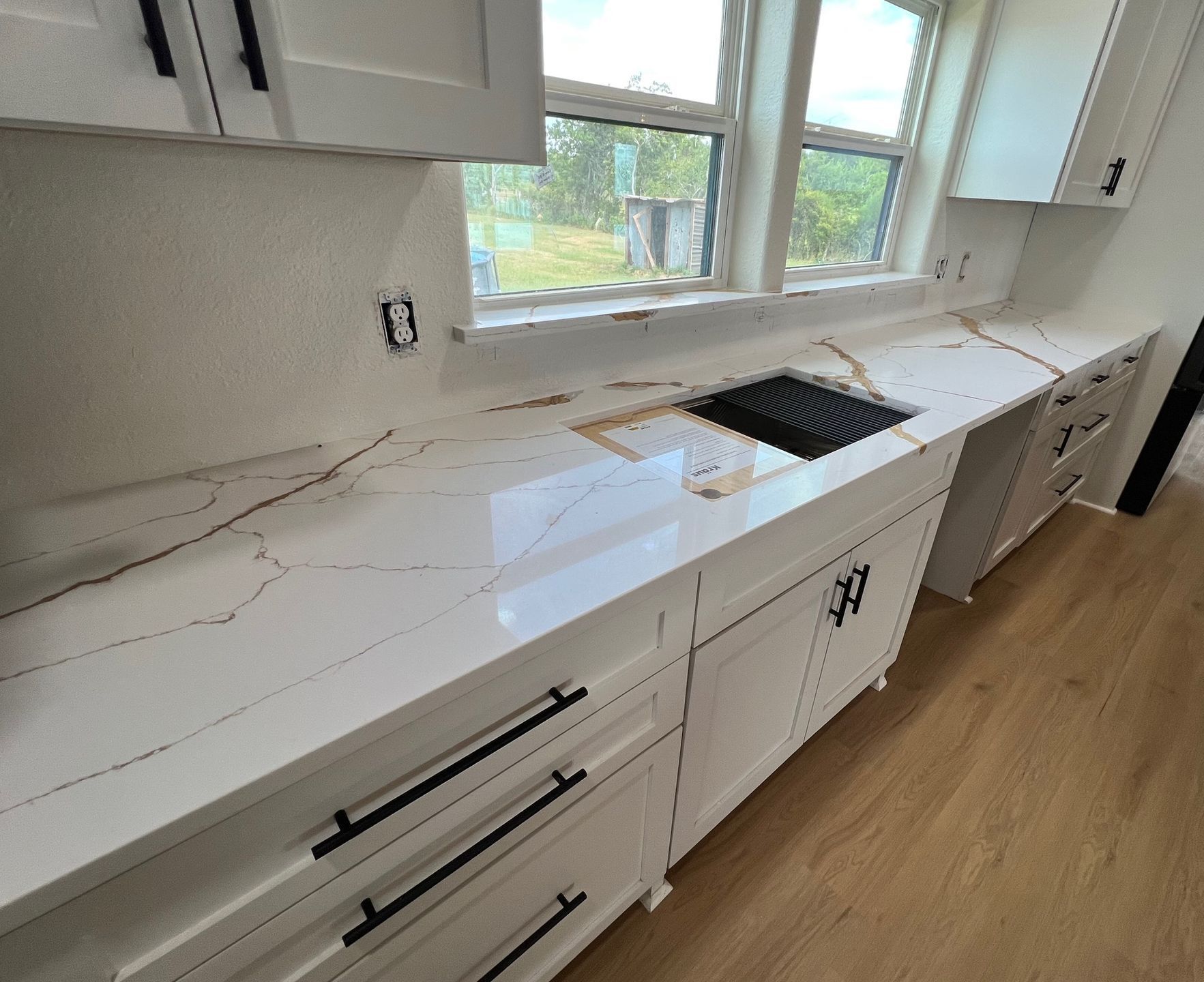 Modern white kitchen with marble-look countertops, farmhouse sink by window, and light wood flooring