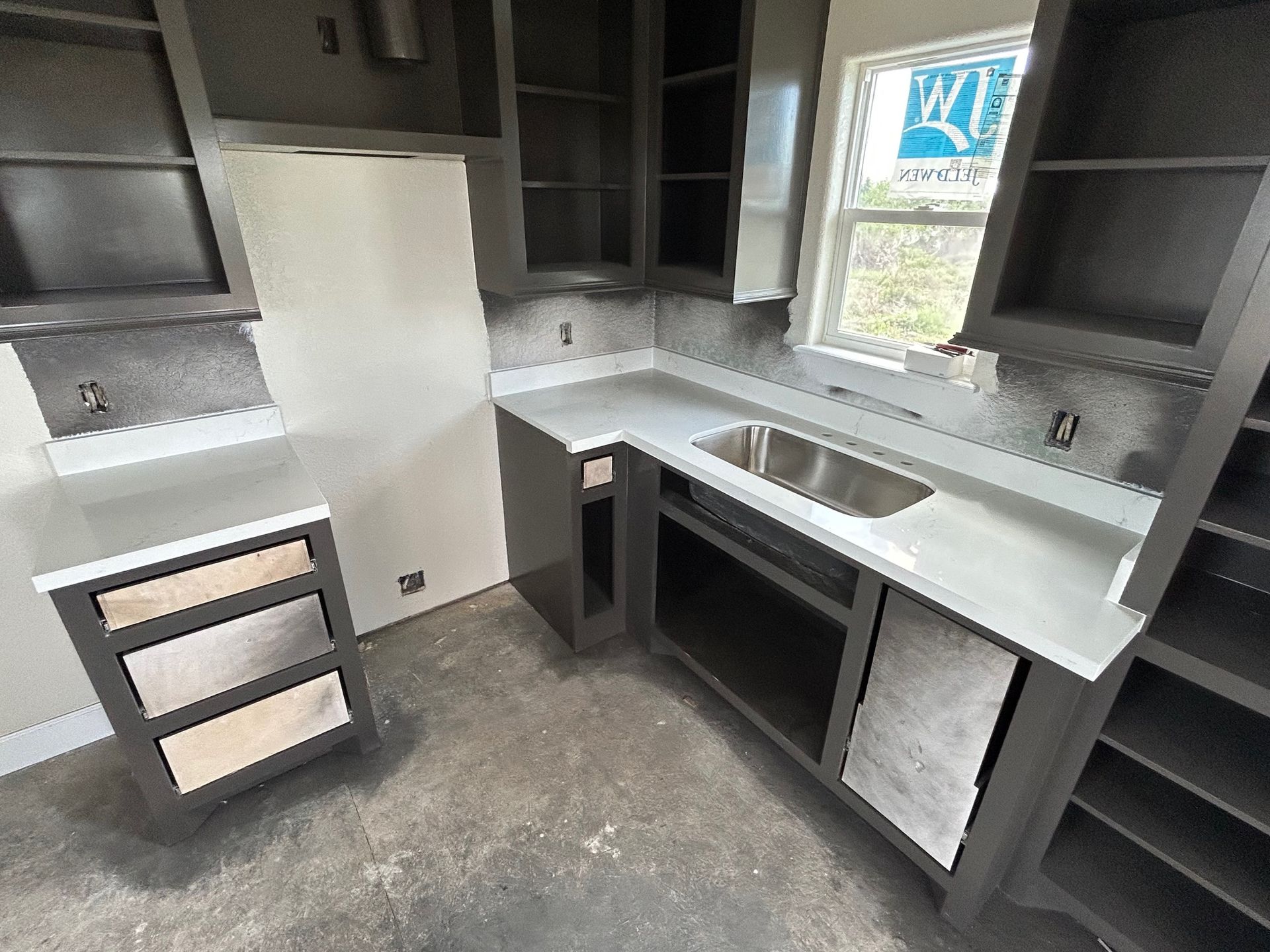 Empty kitchen with dark cabinets, white countertops, a sink by the window, and unfinished lower cabinetry.