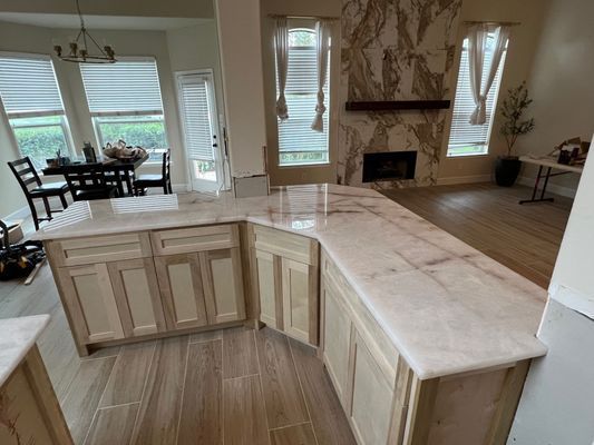 Open kitchen with U-shaped light stone island and cream cabinets, overlooking a bright dining and living area