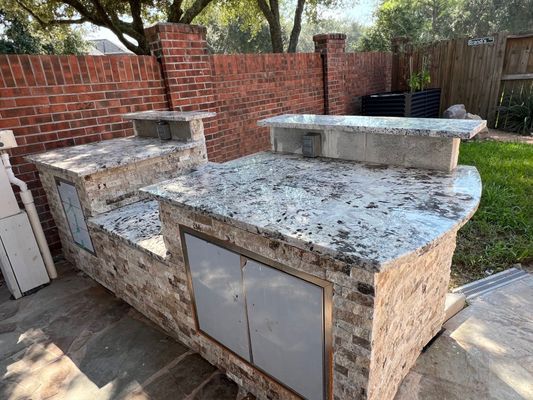 Outdoor stone grill and countertop with metal cabinet doors against a red brick wall in a backyard