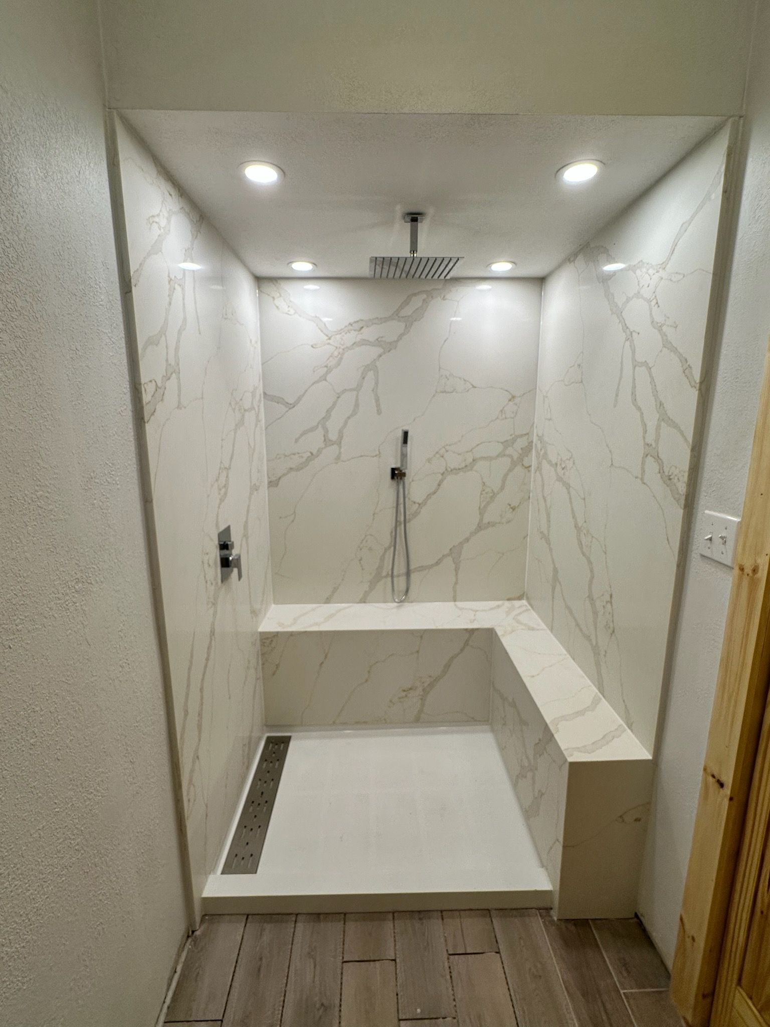 Modern tiled shower with built-in bench, recessed lights, and wood-look floor outside the stall