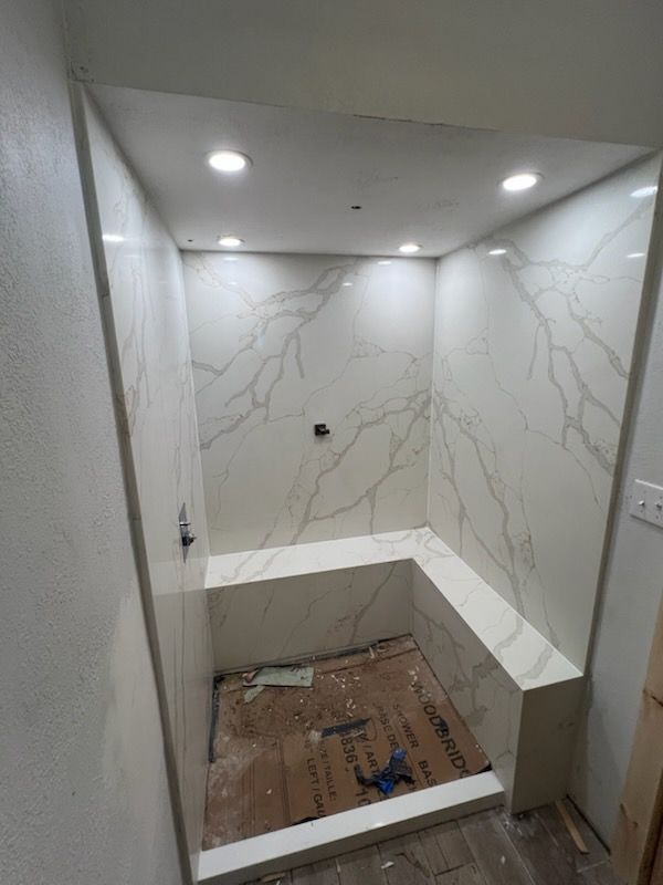 Small tiled shower alcove with built-in bench, marble-look walls, and unfinished floor under recessed lights