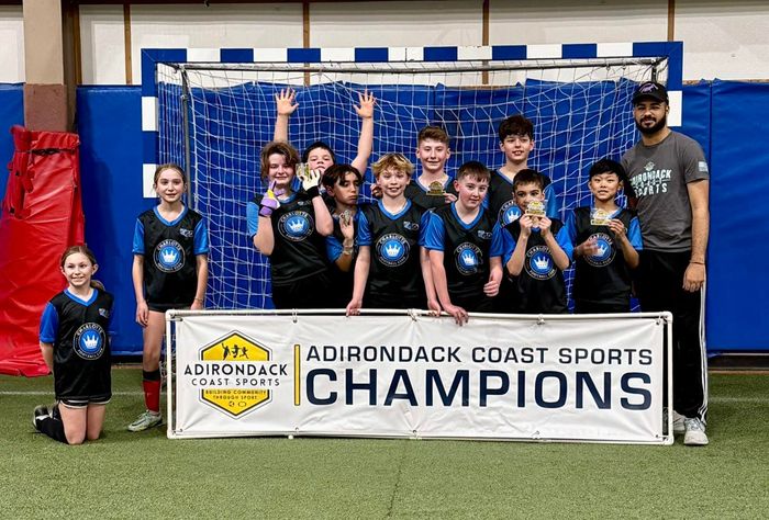 Youth soccer team holding a “Champions” banner in an indoor gym after an Adirondack Coast Sports win.