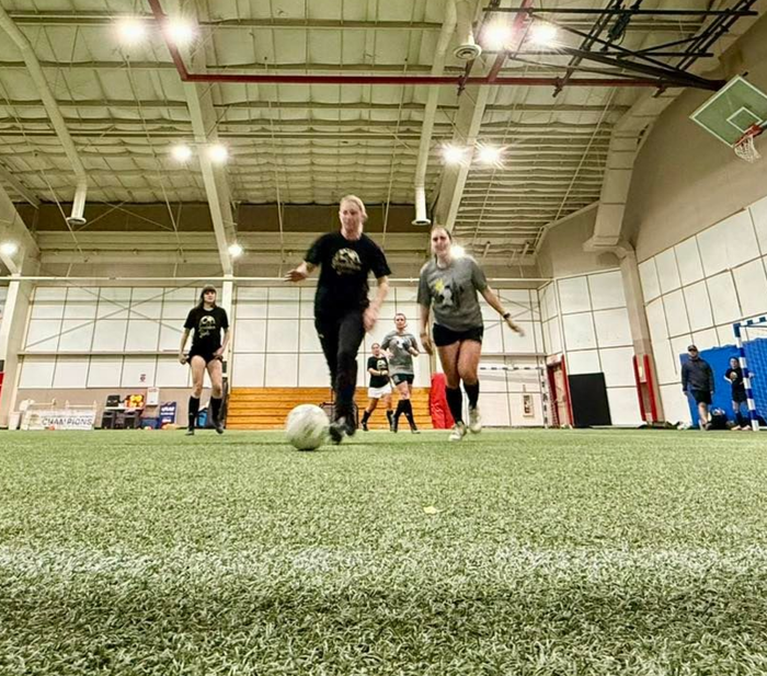 People running toward a soccer ball on an indoor turf field under bright ceiling lights