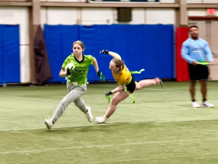 Two athletes sprinting indoors, one in green leading as another in yellow stumbles on turf