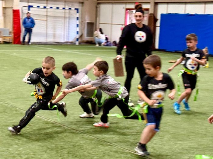 Children sprinting in an indoor soccer game on turf, with a coach watching nearby.