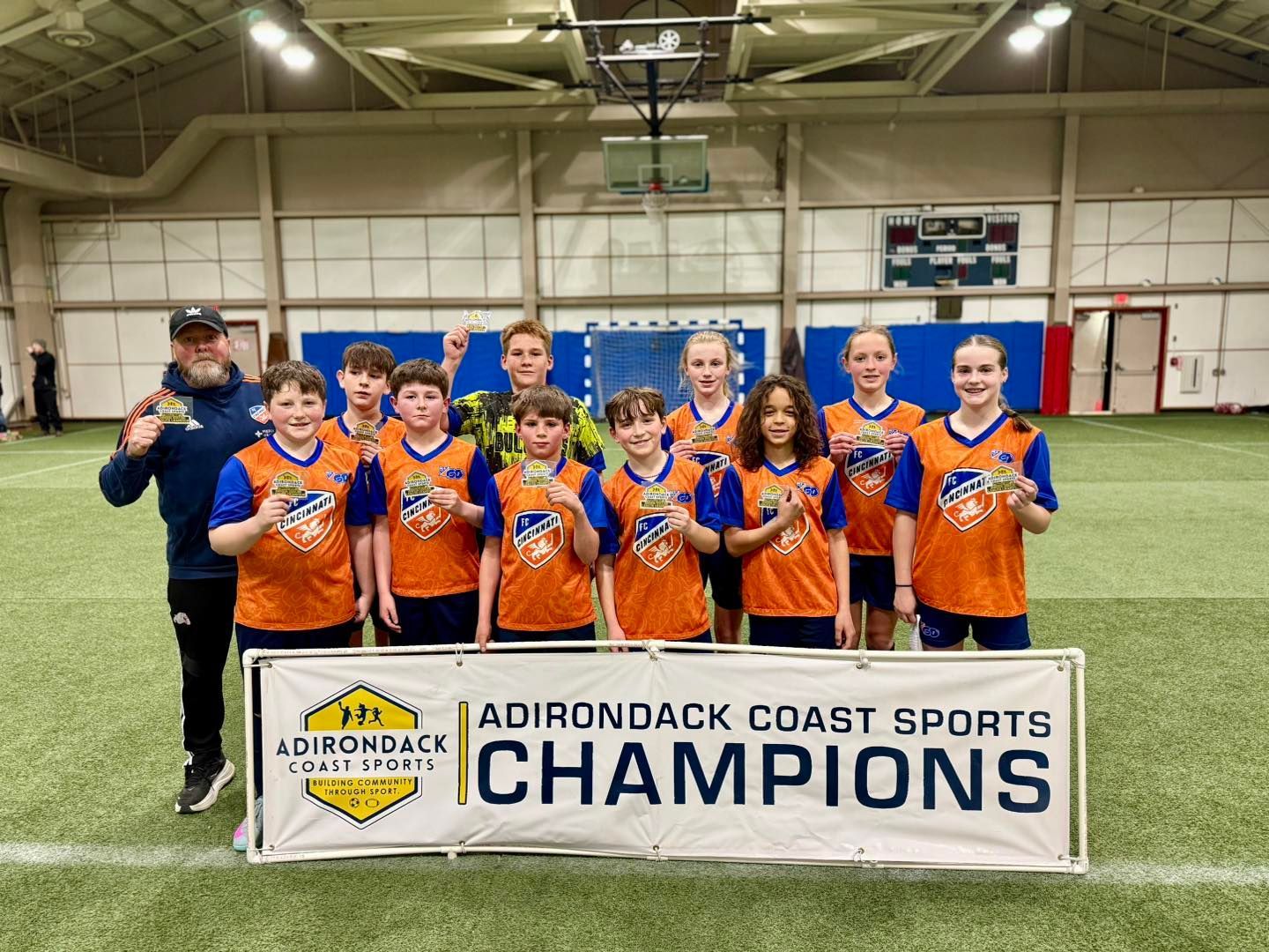 Youth soccer team in blue and orange jerseys celebrating behind a Champions banner indoors