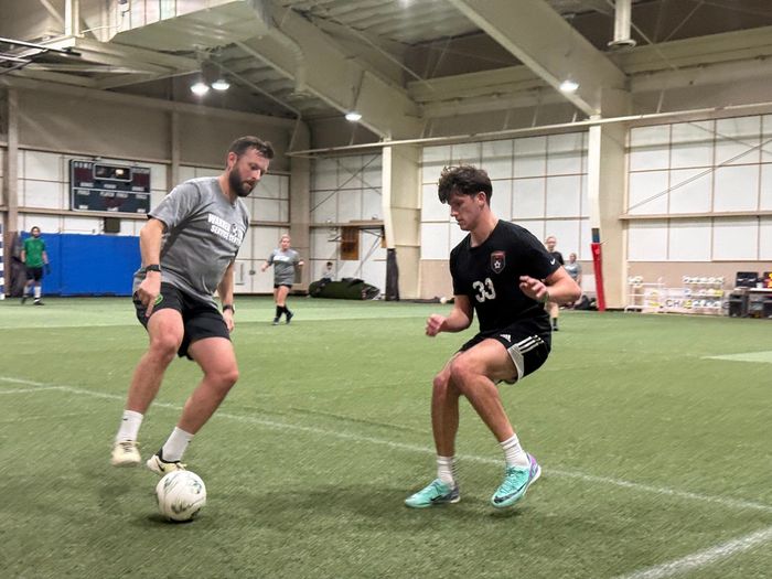 Two athletes dribble soccer balls during an indoor training drill on turf.