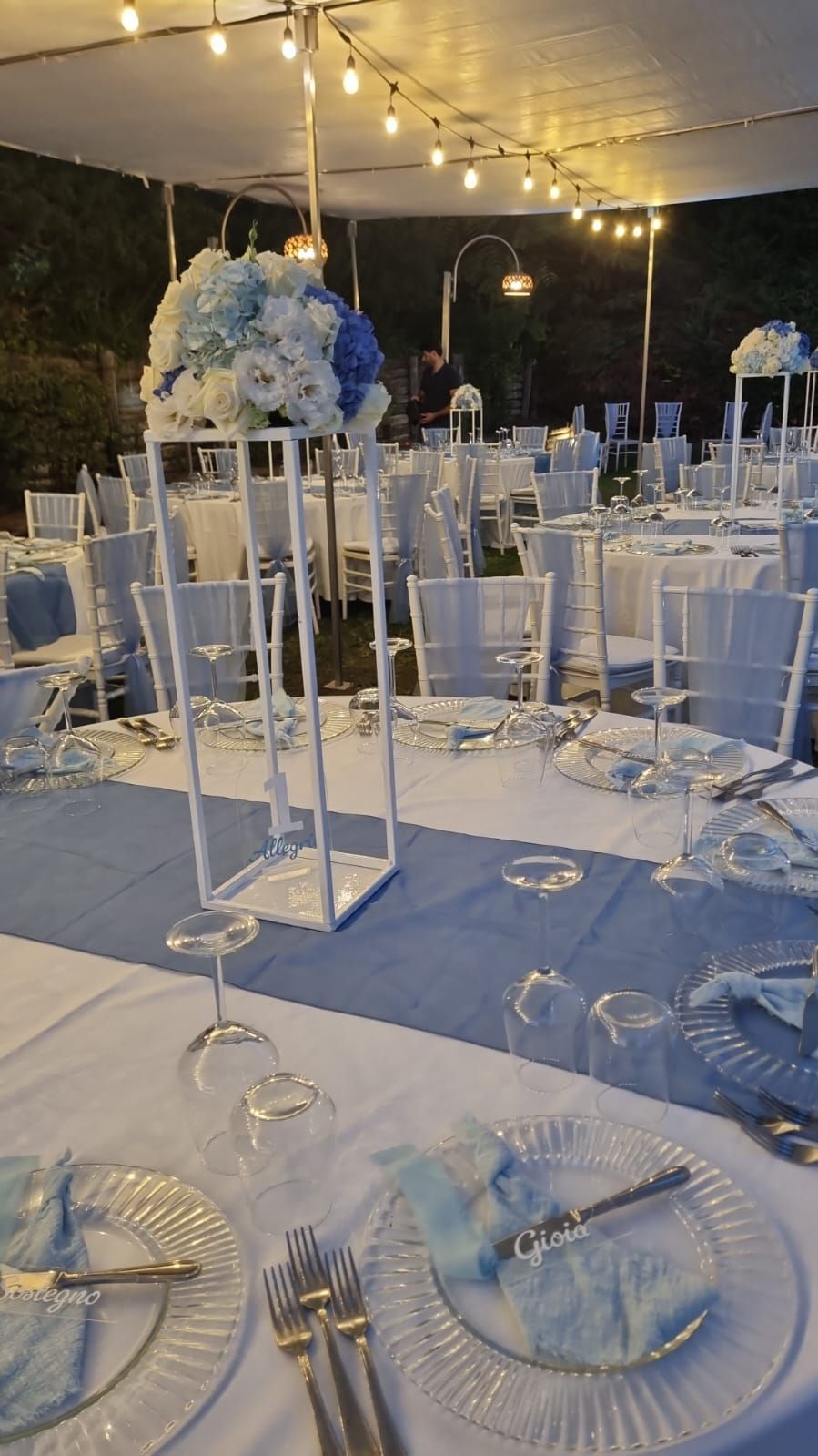 Elegante ricevimento all'aperto con sedie bianche, dettagli blu e centrotavola floreali.