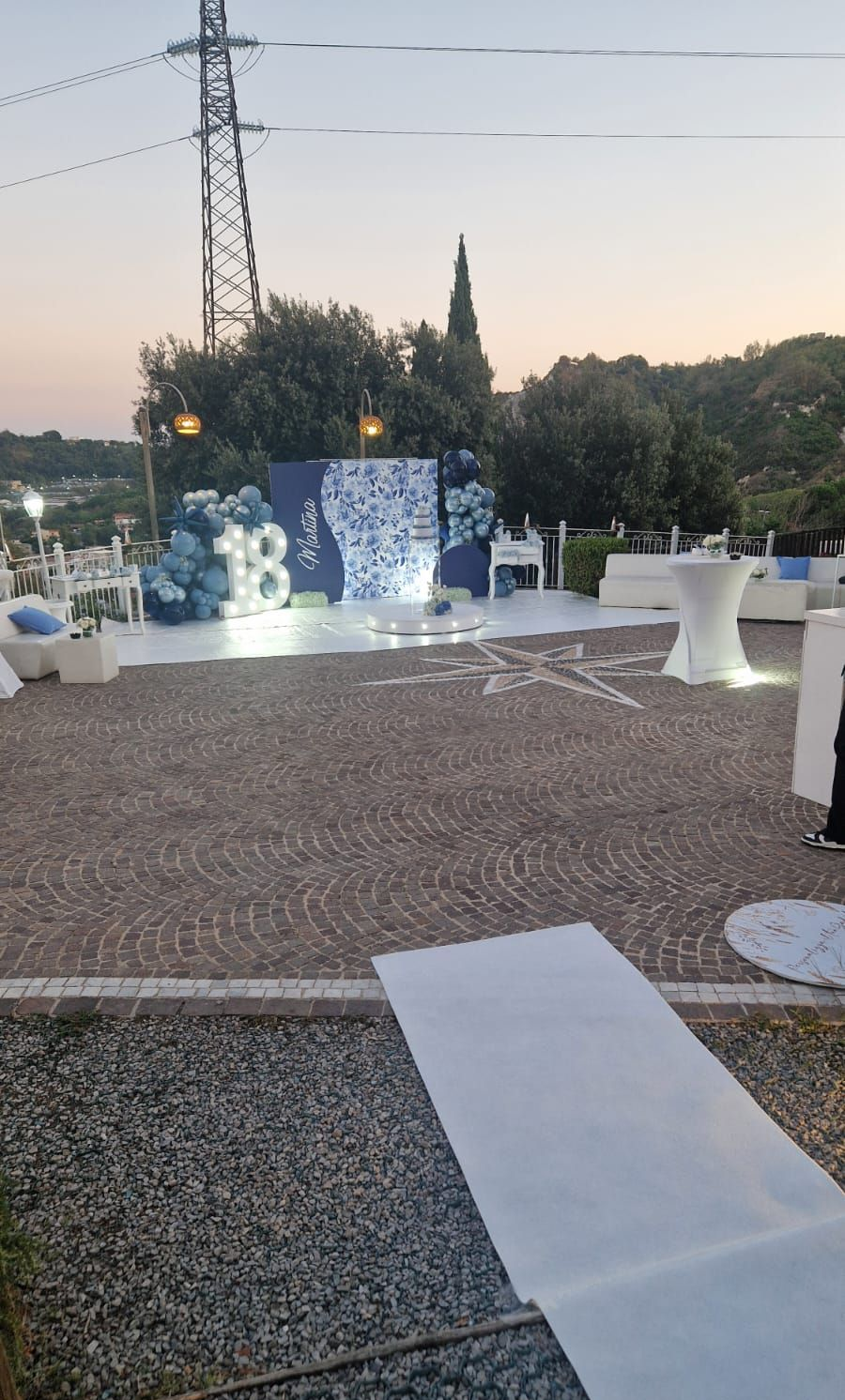 Allestimento per evento all'aperto con decorazioni blu e bianche, tra cui uno sfondo, palloncini e una passerella.