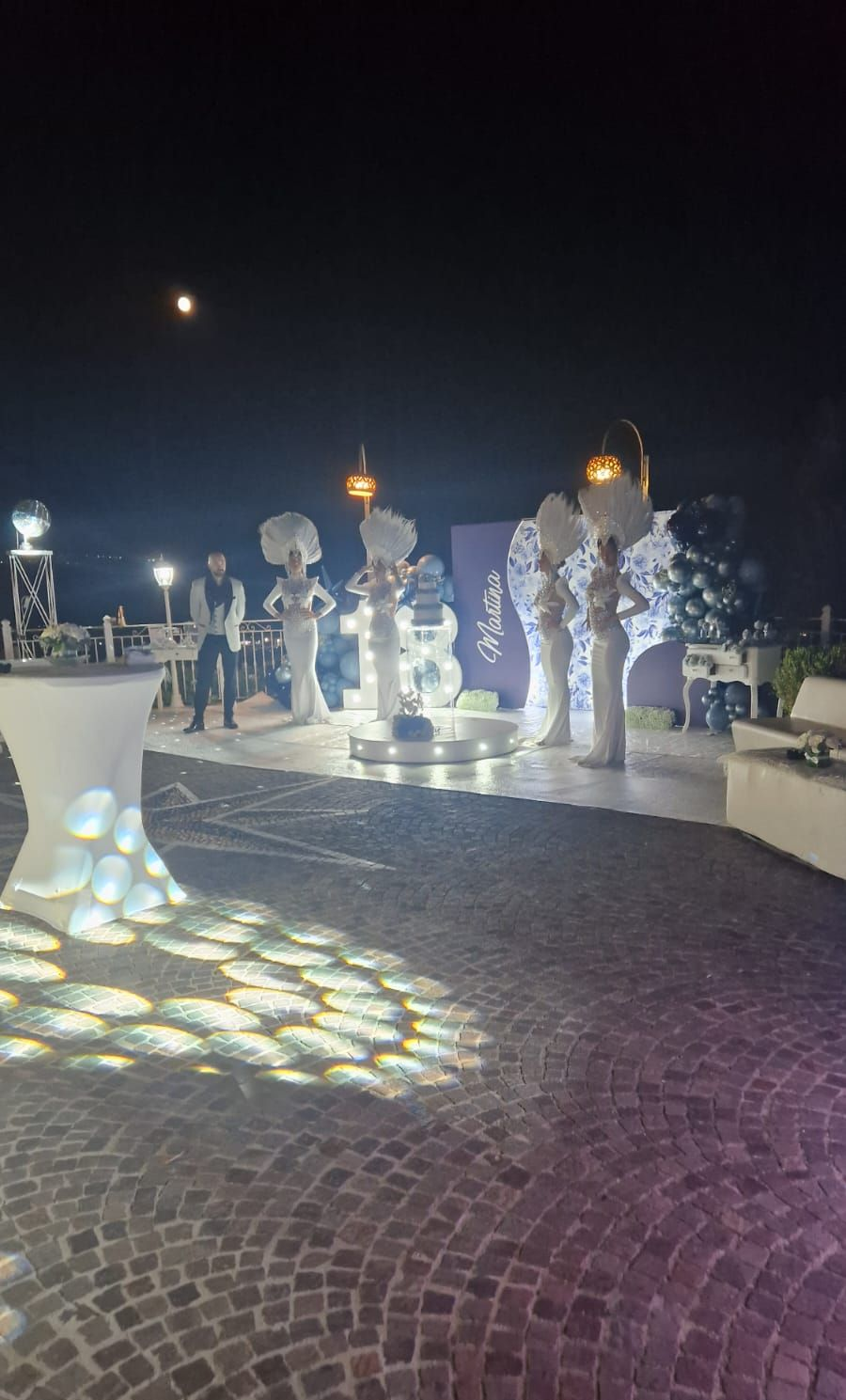 Evento all'aperto di notte con figure e decorazioni illuminate. Luci intense su un patio acciottolato.