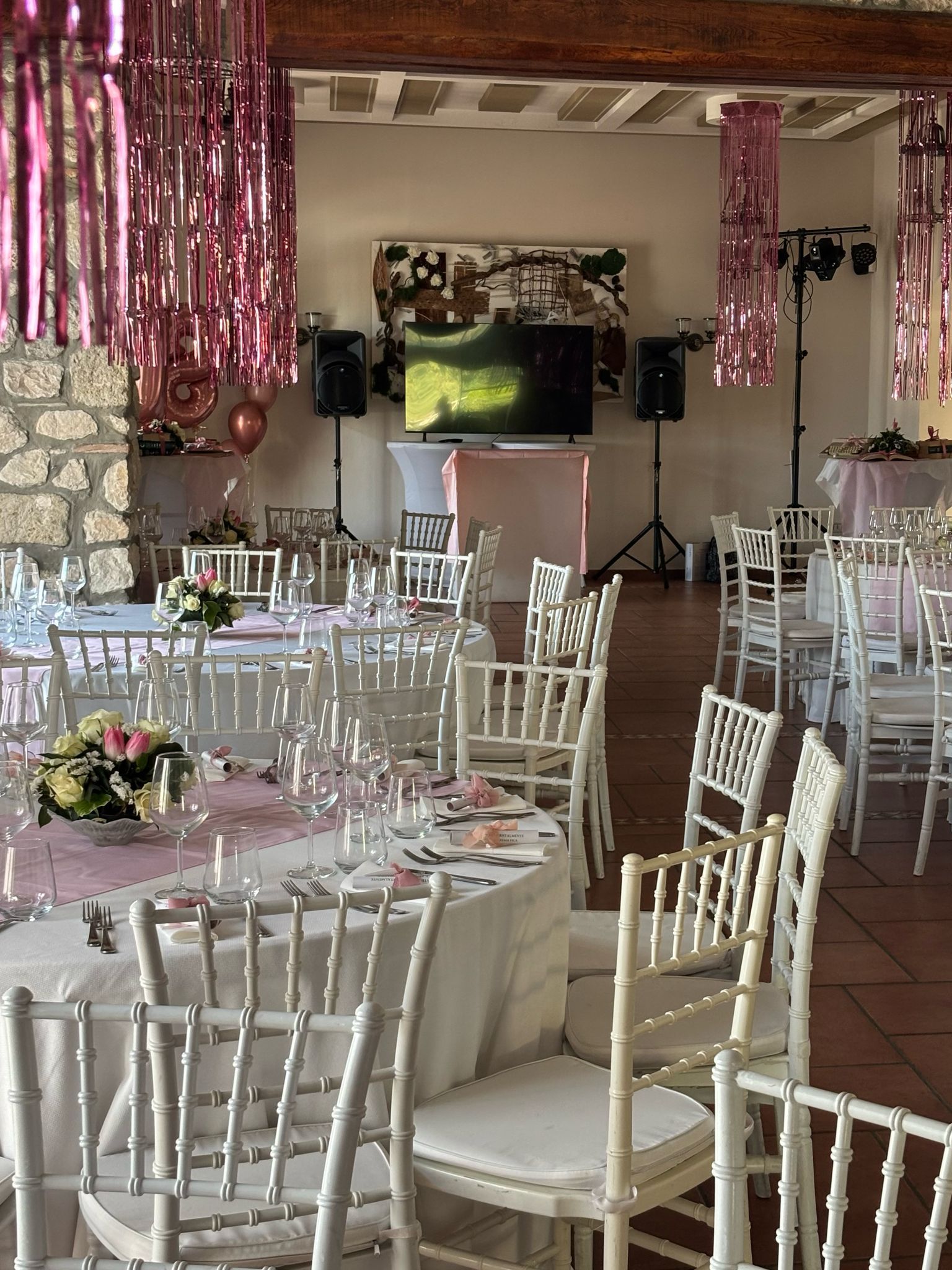 Vista interna di uno spazio per eventi decorato con tavoli e sedie bianchi, decorazioni rosa e una TV.