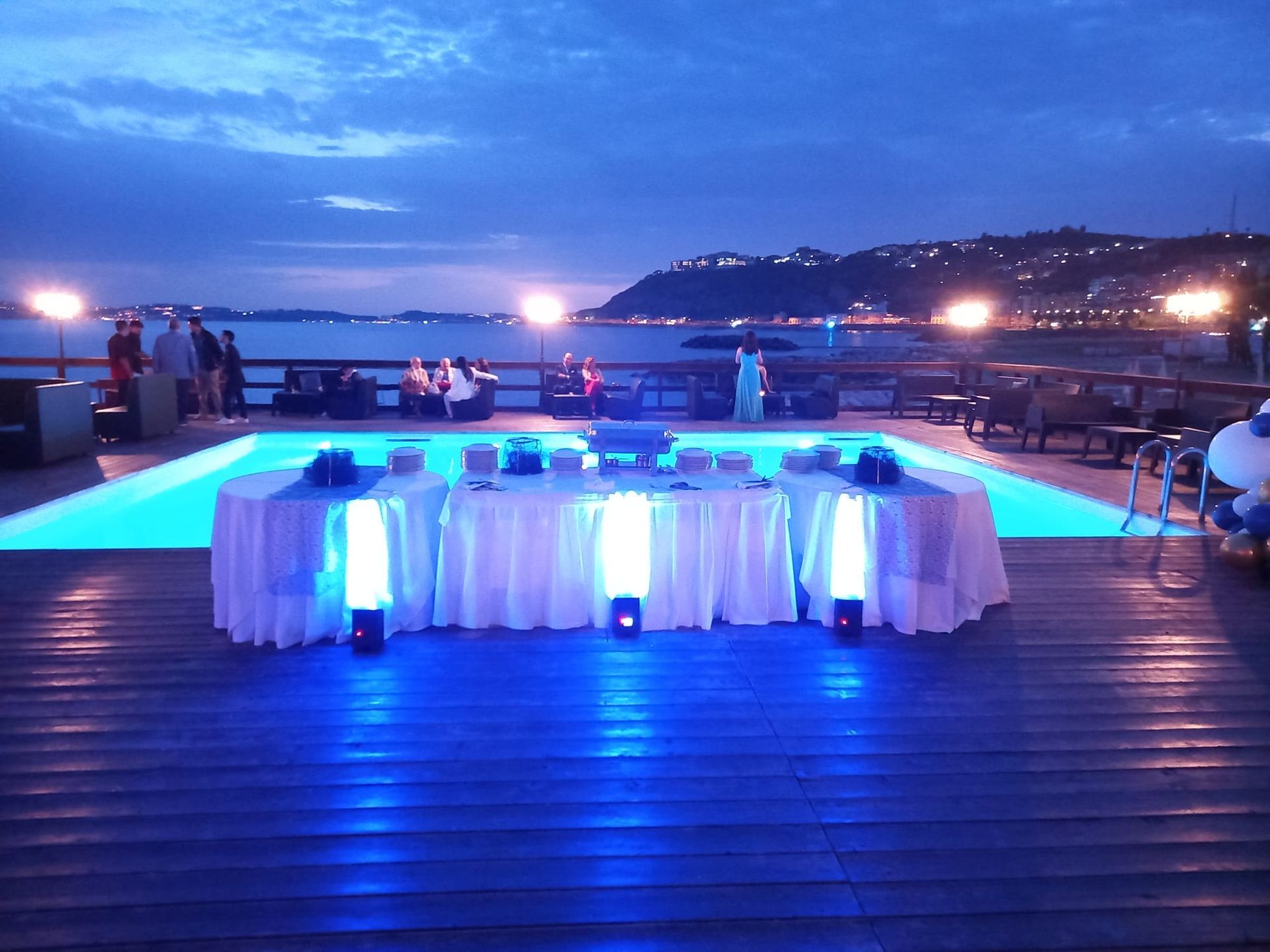 Tavolo apparecchiato per un evento accanto a una piscina blu brillante con acqua e luci. Persone sullo sfondo.