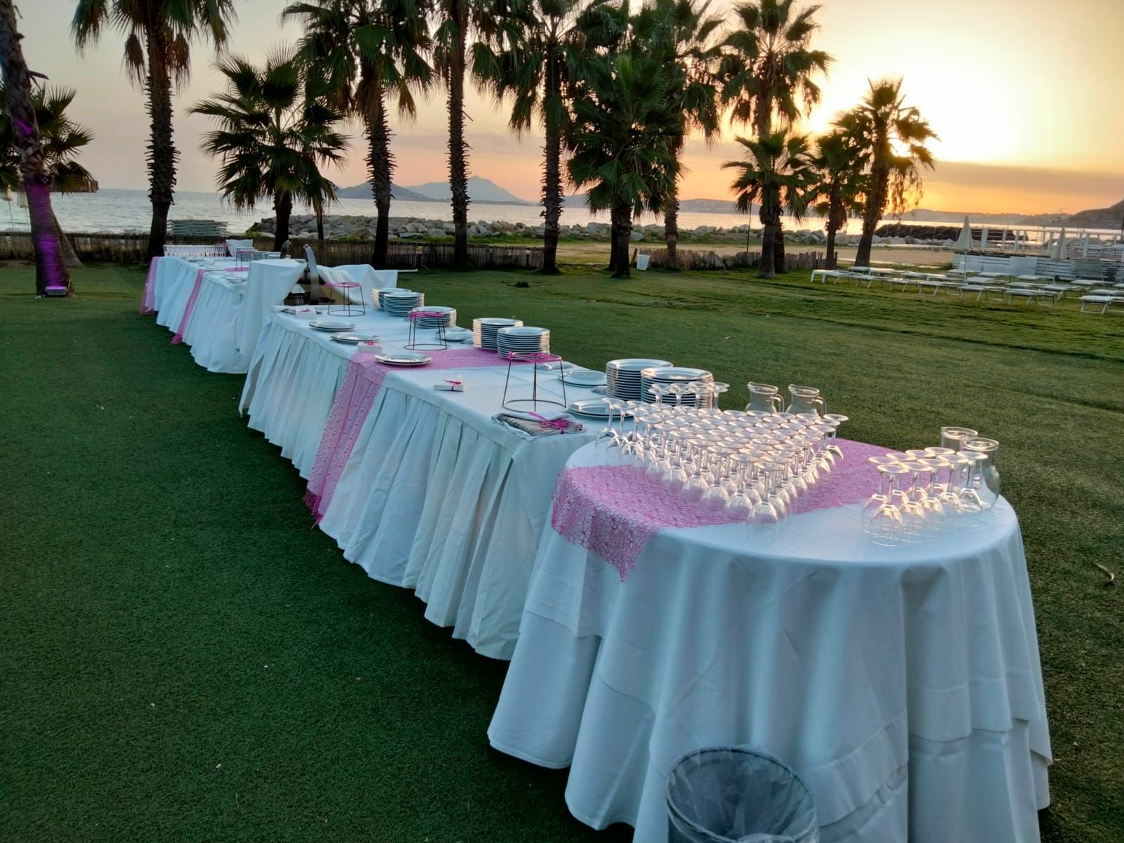 Lunghi tavoli da buffet allestiti all'aperto con tovaglie bianche, dettagli rosa e bicchieri da champagne. 