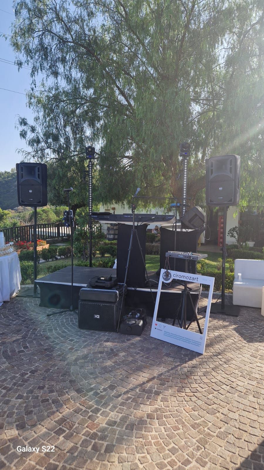 Allestimento di un palco all'aperto con altoparlanti, microfoni e attrezzature su un patio con uno sfondo alberato.