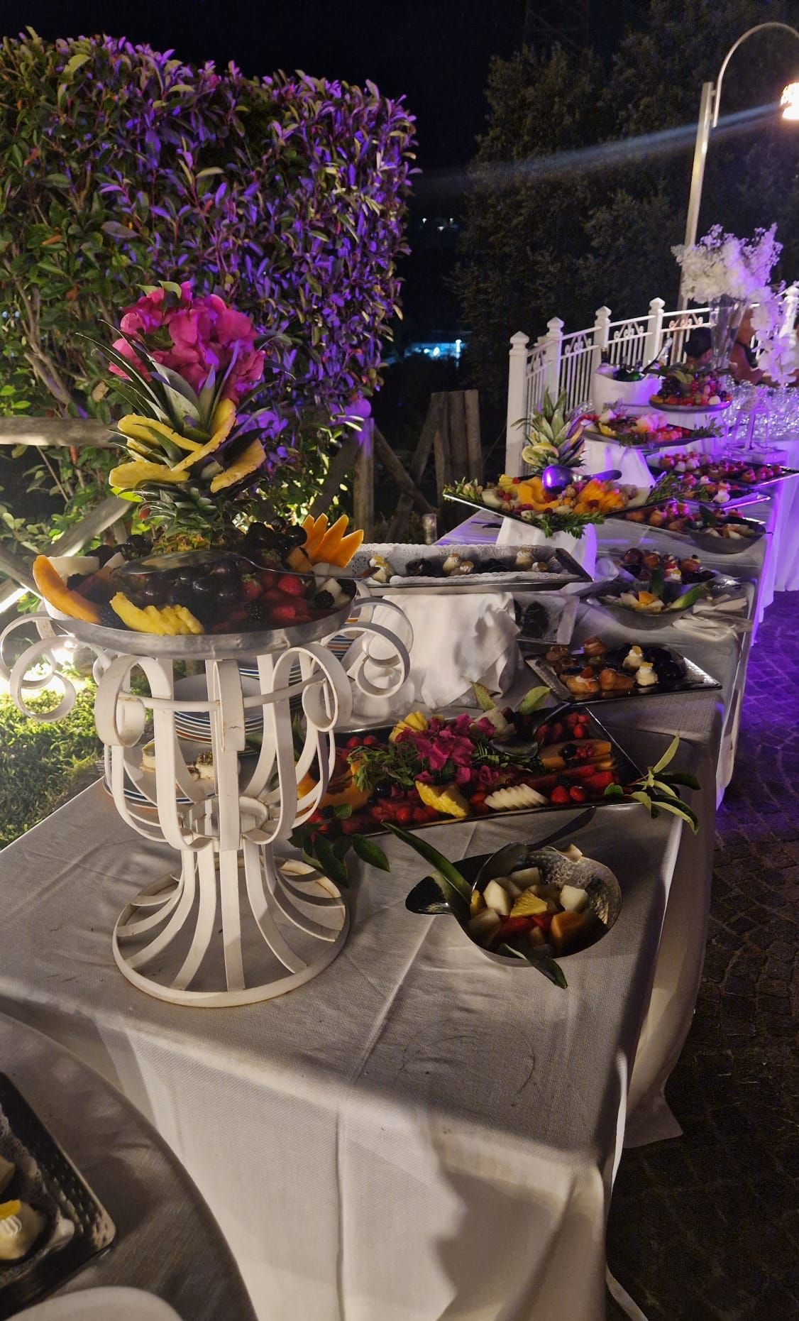 Tavolo da buffet serale, con una varietà di frutta e dessert, disposti in modo decorativo con composizioni floreali.
