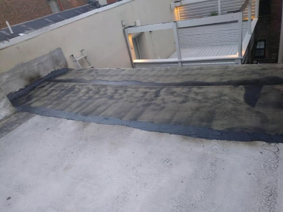 Rooftop with a dark tarp or waterproof strip laid across a light concrete surface near a small gate and wall.